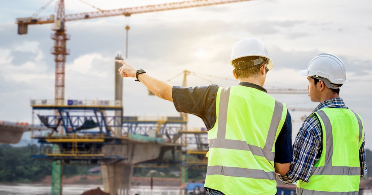 Two construction workers in hard hats stand in front of a bustling construction site, overseeing ongoing work activities.